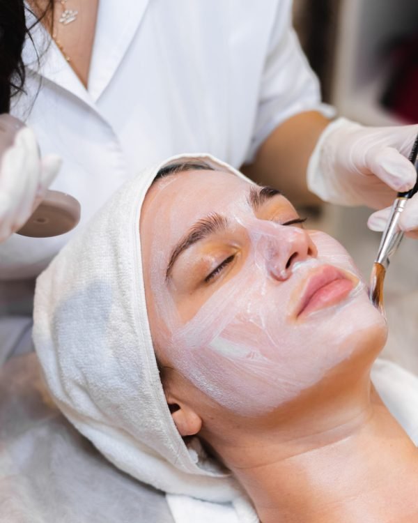 Beautician with a brush applies a white moisturizing mask to the face of a young girl client in a spa beauty salon